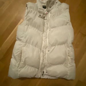 Vit dunväst från Ralph Lauren - Säljer en snygg vit dunväst från Ralph Lauren. Västen har en hög krage och stängs med både dragkedja och knappar. Perfekt för kyliga dagar när du vill hålla värmen utan att kompromissa med stilen.