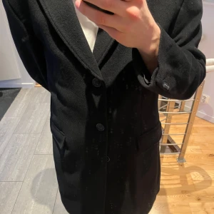 Svart kashmir kappa från Dressmann - Elegant svart kappa från Dressmann i en lyxig kashmir- och ullblandning. Perfekt för att hålla stilen under kyligare dagar. Kappan har en klassisk design med knappar framtill och fickor på sidorna.