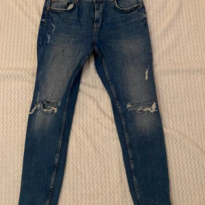 Blå jeans från Bershka - Säljer ett par blå jeans från Bershka med slitna detaljer på knäna och fransiga benslut. Perfekta för en trendig look. Tillverkade i Turkiet.
