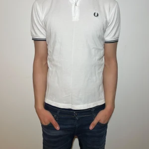 Vit pikétröja från Fred Perry - Snygg vit pikétröja från Fred Perry med korta ärmar och klassisk krage. Tröjan har en broderad logga på bröstet och svarta detaljer vid ärmslut och krage. Perfekt för en stilren look!