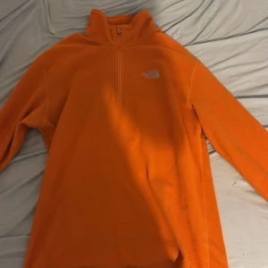 Orange fleece tröja från The North Face - Säljer en orange fleece tröja från The North Face med halv dragkedja och hög krage. Perfekt för kyliga dagar och utomhusaktiviteter. Tröjan har långa ärmar och är supermjuk och bekväm.