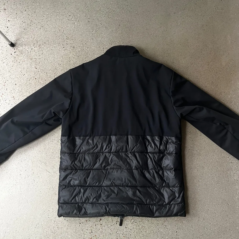 Snygg svart jacka från EA7 med dragkedja och hög krage. Jackan har en quiltad design på nedre delen och är perfekt för kyligare samt regniga dagar. Den har långa ärmar och en stilren look. Passar både S och mindre M.. Takit.