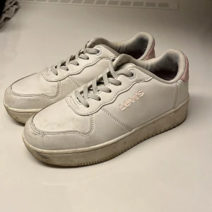 Vita sneakers från Levi's - Snygga vita sneakers från Levi's med rosa detaljer på hälen. Skorna har snörning och en bekväm sula, perfekt för en avslappnad stil. Levi's logga syns tydligt på sidan och baksidan av skorna. Det är ett litet skav märke vid hälen