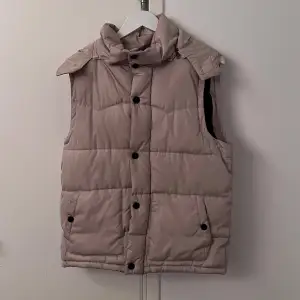 Snygg beige dunväst med avtagbar huva och praktiska fickor. Västen har både dragkedja och knappar för extra skydd mot kyla. Perfekt för lager-på-lager under kyliga dagar. En knapp borta.