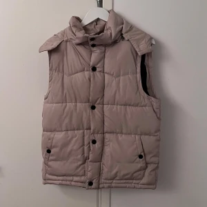 Beige dunväst med huva - Snygg beige dunväst med avtagbar huva och praktiska fickor. Västen har både dragkedja och knappar för extra skydd mot kyla. Perfekt för lager-på-lager under kyliga dagar. En knapp borta.