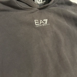 Grå hoodie från Emporio Armani - Säljer en stilren grå hoodie från Emporio Armani med EA7-logga på bröstet och ärmen. Perfekt för en avslappnad look med en touch av lyx. Passar både till vardags och chill. Vid frågor hör av er