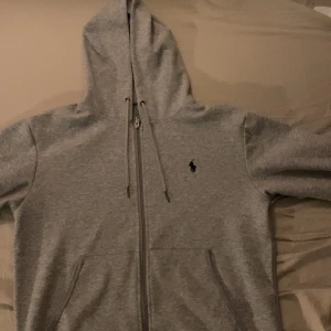 Grå hoodie från Ralph Lauren - Säljer en stilren grå hoodie från Ralph Lauren med dragkedja och klassisk logga på bröstet. Perfekt för en avslappnad look. Den har en justerbar huva och praktiska fickor framtill.
