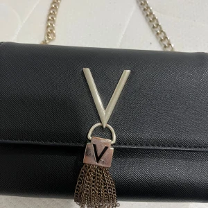 Svart axelväska med kedja - Snygg svart axelväska med en elegant kedja och en dekorativ tofs. Väskan har en stilren design med ett metalliskt V på framsidan. Perfekt för att ge din outfit en touch av glamour. Färgen har försvunnit lite men inget som tar ifrån väskans stilighet.