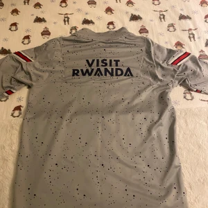Grå fotbollströja från Jordan x PSG - Snygg grå fotbollströja från Jordan i samarbete med PSG. Tröjan har ett coolt stänkmönster och texten 'Visit Rwanda' på ryggen. Framtill finns PSG-loggan och 'Accor Live Limitless'. Ärmarna har röd och svart randdetalj sälj för 450kr jätte bra aldrig nästan använd hör av er för mer frågor 
