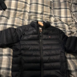 Mörk blå dunjacka från Tommy Hilfiger - Säljer en stilren svart dunjacka från Tommy Hilfiger. Jackan har en quiltad design med dragkedja framtill och en liten logotyp på bröstet. Perfekt för kyligare dagar.  Kan gå ner på priset vid en snabb affär