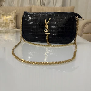 Svart axelväska med kedja - Elegant svart axelväska med krokodilpräglat mönster och guldfärgad kedja. Väskan har en dragkedja upptill och en stilren design med en logotypdetalj framtill.