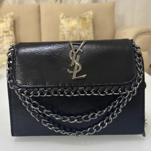 Svart axelväska med kedjedetaljer - Snygg svart axelväska från Yves Saint Laurent med kedjedetaljer och logotyp på framsidan. Väskan har en stilren design med flera dragkedjor och en justerbar kedjerem. Perfekt för att ge en lyxig touch till din outfit.