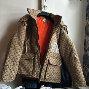 Beige mönstrad jacka från The North Face x Gucci - Säljer en unik beige jacka med ikoniskt mönster från The North Face x Gucci. Jackan har en orange insida, stora fickor med knappar och en praktisk huva. Perfekt för att sticka ut i mängden! Uppskattad storlek M, köpt second hand men väldigt fint skick! 