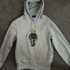 Grå hoodie från Polo Ralph Lauren - Säljer en grå hoodie från Polo Ralph Lauren med en ikonisk nallebjörn på framsidan. Tröjan har en justerbar huva med dragsko och ribbade muddar vid ärmslut och nederkant. Perfekt för en avslappnad stil. Nypris: 2700