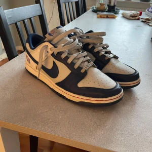 Nike Dunk Low med Brooklyn Nets tema - Snygga Nike Dunk Low sneakers i vitt och blått med Brooklyn Nets logga på hälen. Skorna har klassisk snörning och en stilren design som passar perfekt för alla sneakerheads.