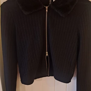 Svart ribbad jacka med fuskpäls - Snygg svart ribbad jacka med dragkedja och mjuk fuskpäls på kragen och ärmsluten. Dragkedja som kan öppnas även nederkant. Perfekt för kyliga dagar när du vill vara både varm och stilren.