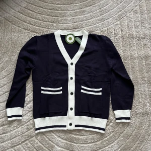 Mörkblå Cardigan HELT NYTT - HELT NY OCH OANVÄND!! Snygg mörkblå Cardigan med vita kanter och knappar. Två fickor framtill med vita ränder ger en stilren look. Perfekt för en avslappnad men ändå elegant stil.