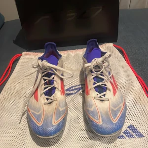 Adidas F50 elite Ag - Snygga Adidas fotbollsskor i vitt med blå och röda detaljer. Skorna har snörning och en modern design som passar perfekt för fotbollsplanen. De har en strömlinjeformad form och är gjorda för att ge bra grepp och komfort.