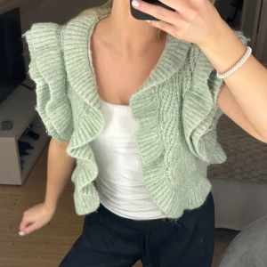 Mintgrön stickad kofta med volangdetaljer - Säljer en söt mintgrön stickad kofta med volangdetaljer. Koftan är kortärmad och har en öppen design som ger en luftig känsla. Perfekt för lager-på-lager-stil och att piffa upp vilken outfit som helst.