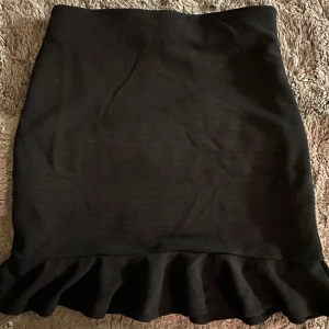 Svart kort kjol med volang - Snygg svart kjol med volangdetalj i nederkant. Perfekt för en stilren look. Kjolen har en tight passform och är gjord i ett stretchigt material för extra komfort. 