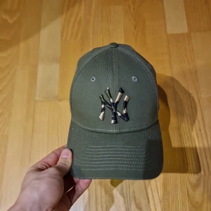 Grön keps från New Era - Snygg grön keps från New Era med broderad logotyp i camouflage på framsidan. Kepsen är tillverkad i 100% bomull och har en klassisk passform med justerbar rem baktill.