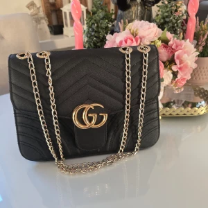 Svart axelväska med silverkedja - Snygg svart axelväska i skinn med elegant silver och GG-logga framtill. Väskan har ett quiltat mönster och stängs med en tryckknapp. Perfekt för att ge en lyxig touch till din outfit.