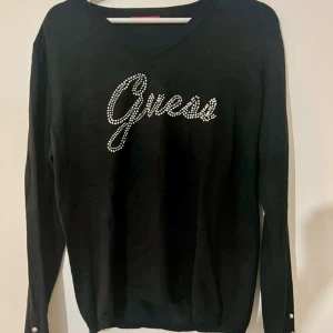 Svart tröja från Guess med pärlor - Snygg svart tröja från Guess med pärldetaljer som bildar loggan på framsidan. Tröjan har långa ärmar och dekorativa knappar vid ärmsluten. Perfekt för en stilren look.