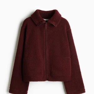 Vinröd teddyjacka med dragkedja - Mysig vinröd teddyjacka med dragkedja framtill och två fickor. Perfekt för kyliga dagar när du vill hålla dig varm och bekväm. Helt ny med prislappen kvar