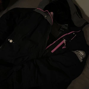 Svart jacka med rosa detaljer från Peak Performance  - jacka från Peak Performance med rosa dragkedjor och detaljer. Jackan har en huva med grå insida och en logga på ärmen. Perfekt för kyliga dagar.