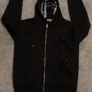 Svart huvtröja från Burberry - Säljer en stilren svart huvtröja från Burberry i storlek M . Det är en klassisk zip hoodie med dragkedja och klassiskt rutigt mönster i fodret. Tröjan har långa ärmar och en bekväm passform. Perfekt för en avslappnad look.
