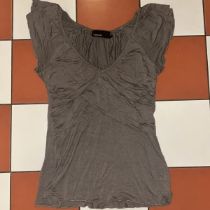 Grå topp från KappAhl - Säljer en stilren grå topp från KappAhl med korta ärmar och v-ringad design. Perfekt för en avslappnad look. Toppen har en enkel och bekväm passform.