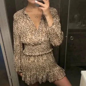 Leopardmönstrad klänning - Trendig leopardmönstrad klänning med volangdetaljer och långa ärmar. Klänningen har en v-ringad hals och en smickrande passform som markerar midjan. Perfekt för en stilfull look.