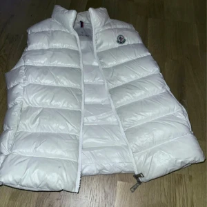 Vit dunväst från Moncler - Säljer en snygg vit dunväst från Moncler med dragkedja och ikonisk logga på bröstet. Perfekt för kyliga dagar när du vill hålla stilen. Västen har en quiltad design som ger extra värme och komfort.