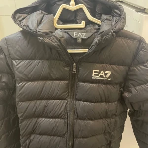 Emporio Armani jacka - Snygg svart dunjacka från Emporio Armani EA7 med huva och dragkedja. Perfekt för kyligare dagar. Storlek 175cm. Jag är tjej på 165cm oxh den passar mig. Den passar även mina två bröder som är 170 och 180cm långa. Perfekt jacka för båda könen! Den är gjord för män men ser helt otrolig fin ut på tjejer. Köptes denna vintern förra månaden.