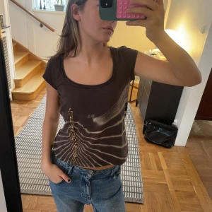 Brun kortärmad topp med mönster - Snygg brun kortärmad topp med unikt mönster och dekorativa detaljer framtill. Perfekt för en avslappnad stil.