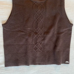 Brun stickad väst  - Säljer en brun stickad väst med ett snyggt flätmönster framtill. Västen har ribbad nederkant och är perfekt för lager-på-lager-stil. Passar bra till både skjorta och t-shirt.
