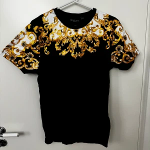 Svart t-shirt med guldmönster strl M - Snygg svart t-shirt från Brave Soul med ett iögonfallande guldmönster över axlar och bröst. Perfekt för att sticka ut i mängden. Kortärmad och tillverkad i mjukt material för en bekväm passform. Aldrig använd. Storlek M
