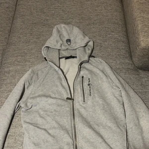 Grå huvtröja från Sail racing - Säljer en grå huvtröja från sail racing med dragkedja och tryckt logga på bröstet. Tröjan har en justerbar huva och långa ärmar, perfekt för en avslappnad stil.