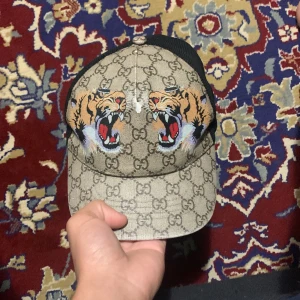 Beige keps med tigerbroderi - Snygg beige keps med svart nät och broderade tigrar framtill. Kepsen har ett klassiskt mönster och justerbar passform. Perfekt för att ge din outfit en cool touch!