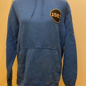 Blå hoodie S 1987 - Snygg blå hoodie med ett stort 1987-märke på bröstet. Tillverkad i en mjuk blandning av bomull och polyester. Perfekt för en avslappnad stil med känguruficka och justerbar huva.  Armhåla till armhåla: 55cm 
