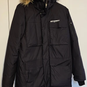 Svart parkajacka från Everest - Snygg svart parkajacka från Everest med pälskantad huva. Jackan har flera praktiska fickor och en dragkedja framtill. Perfekt för kalla dagar.