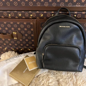 Svart ryggsäck från Michael Kors - Snygg svart ryggsäck i skinn från Michael Kors med guldfärgade detaljer och dragkedja. Perfekt för att bära dina dagliga nödvändigheter med stil. Ryggsäcken har justerbara axelremmar och ett praktiskt ytterfack med dragkedja. Använd 3-5 gånger så väldigt bra pris. 