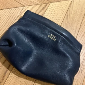 Svart kuvertväska/neccessär  från Acne Studios - Elegant svart kuvertväska i skinn från Acne Studios. Perfekt för att bära dina viktigaste saker med stil. Diskret logotyp i guld på framsidan ger en lyxig touch.