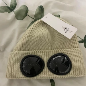 Beige mössa från C.P. Company - Snygg beige stickad mössa från C.P. Company med unika glasögondetaljer framtill. Perfekt för att hålla stilen under kyliga dagar.