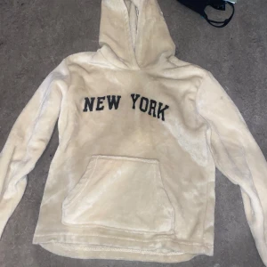 Beige hoodie med texten 'New York' - Mysig beige hoodie med texten 'New York' på bröstet. Tröjan har en stor ficka framtill och en bekväm huva. Perfekt för en avslappnad stil.