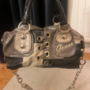 Svart och beige handväska från Guess - Snygg handväska från Guess i svart och beige med gulddetaljer. Väskan har en kedja som axelrem och dekorativa snördetaljer framtill. Väskan har ett tillhörande längre band av kedja och tyg som går att ta av och på. I väskan finns  en ficka med dragkedja och två fack. 
