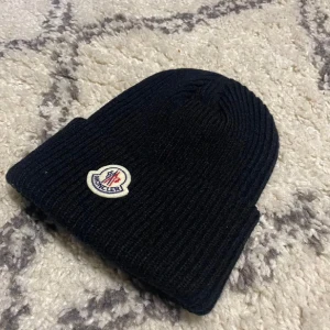 Moncler mössa - Snygg svart ribbad mössa från Moncler med broderad logotyp framtill. Perfekt för kyliga dagar och ger en stilren look. Passar både till vardags och mer uppklädda tillfällen.