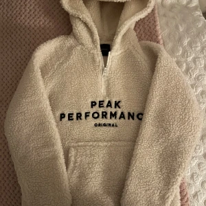 Beige fleece hoodie från Peak Performance - Mysig beige fleece/teddy hoodie från Peak Performance med dragkedja och känguruficka. Perfekt för kyliga dagar med sin mjuka och varma design. Stort tryck på framsidan ger en sportig look. Storlek 140 elr XXXS. Pris går att diskuteras.