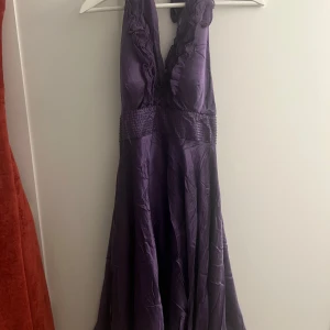 Lila klänning från Adrianna Papell - Elegant lila klänning från Adrianna Papell med volangdetaljer och en v-ringad design. Klänningen har en smickrande passform med ett glittrigt midjeband som ger en extra touch av glamour. Skulle säga att den passar storlek 34-38 då klänningen är justerbar vid halsen. 100% silk. 