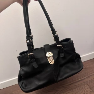 Svart handväska med gulddetaljer - Snygg svart handväska med dubbla handtag och elegant guldfärgad låsdetalj framtill. Perfekt för att bära dina dagliga nödvändigheter med stil.